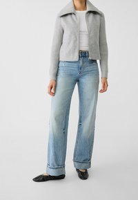 Gray zip-up jacket with a wide collar, ribbed sleeves, light blue wide-leg jeans with distressed detailing, and black flats.