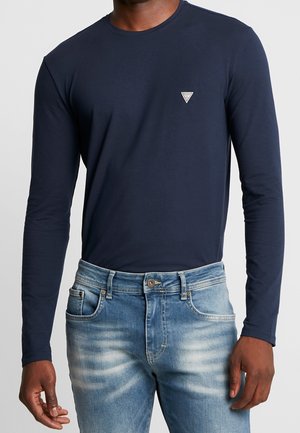 Hombre con camisa de manga larga azul marino con pequeño logo de triángulo en el pecho y vaqueros azules descoloridos, de pie contra un fondo liso.