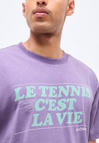 Κοντινό πλάνο ενός ατόμου που φοράει μοβ μπλούζα με πράσινο μέντας κείμενο "LE TENNIS C'EST LA VIE" και εμφανίζεται μερικώς το όνομα της μάρκας "BJÖRN".