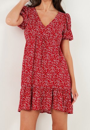 Femme portant une mini robe rouge à manches courtes avec un petit imprimé floral blanc et un col en V, debout devant un fond uni.
