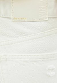 White denim fabric with textured surface. Features a tag labeled "Bershka" and stitching details. Small frayed hole accent visible.