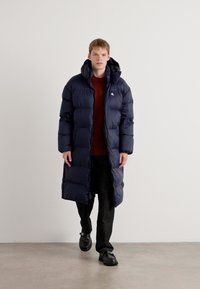 Mørkeblå dunjakke med høy krage, med horisontale quiltede seksjoner og en lang design, kombinert med en maroon topp og svarte bukser.