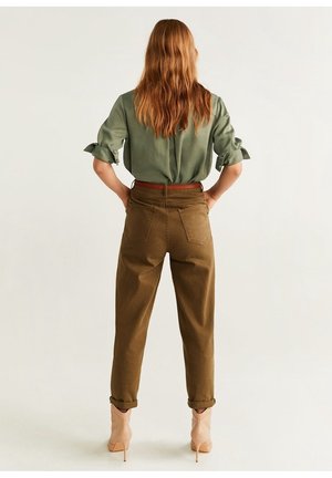 Frau mit langen roten Haaren, stehend, trägt ein hochgekrempeltes olivgrünes Hemd, braune Hose mit hoher Taille, braunen Gürtel und beige Stiefel mit hohen Absätzen.
