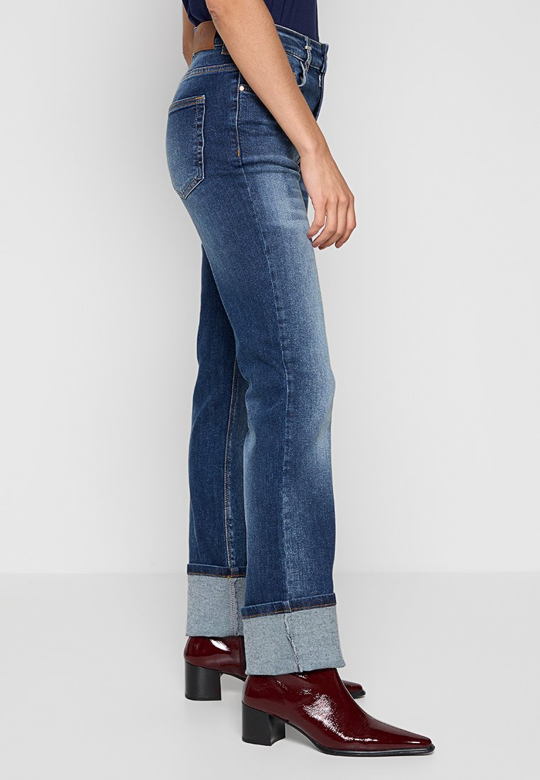 Jasnoniebieskie dżinsy o wysokim stanie, wykonane z denimu z lekko wyblakłym wykończeniem, z podwiniętymi mankietami w jaśniejszym odcieniu denimu, akcentowane błyszczącymi burgundowymi botkami do kostek.