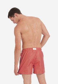 Punaiset uimapuvut joustavalla vyötäröllä, joissa on sivutaskut ja pieni merkkilogo takana, valmistettu kevyestä materiaalista.