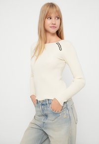 Ραφή κρεμ ζακέτα με τετράγωνη λαιμόκοψη, με μαύρες λεπτομέρειες στους ώμους. Συνδυάζεται με ανοιχτού μπλε, φαρδιά παντελόνια.