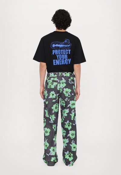 Schwarzes T-Shirt mit blauer Schrift und Illustration auf dem Rücken; graue Blumenhose mit grünen und lila Blumen; lockerer Schnitt.