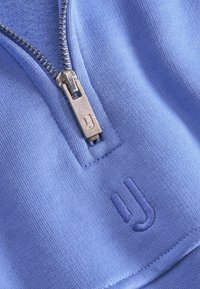 Blauwe sweatshirt met een rits in de hals, voorzien van een zilveren rits en een geborduurd, getextureerd logo op de stof. Over het algemeen glad materiaal.