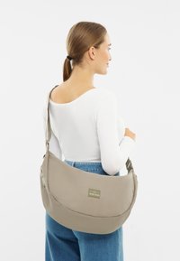 Beige válltáska puha anyagból, lekerekített formával, állítható pánttal és egy zöld logós húzóval az oldalán.