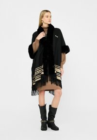 Donna che indossa un poncho nero bordato di pelliccia sopra un vestito di maglia marrone e stivali neri fino al polpaccio con fascette dorate, in piedi su uno sfondo bianco.