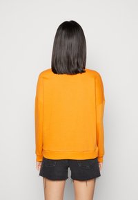 Pullover arancione con maniche lunghe, texture morbida, spalle cadenti e vestibilità rilassata, abbinato a pantaloni corti in denim neri.