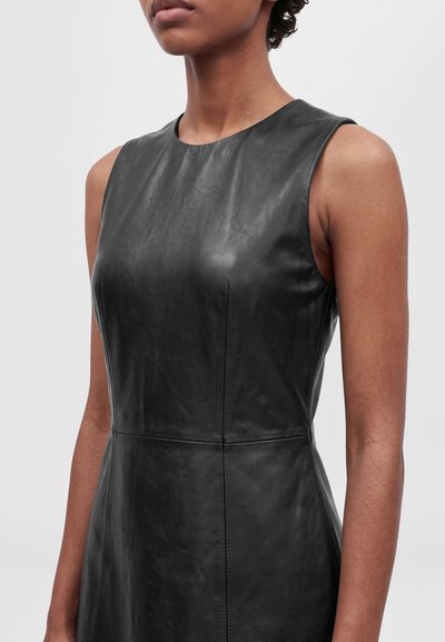 Un mannequin porte une robe en cuir noire ajustée et sans manches, à encolure ronde et avec une couture à la taille, sur un fond clair uni.
