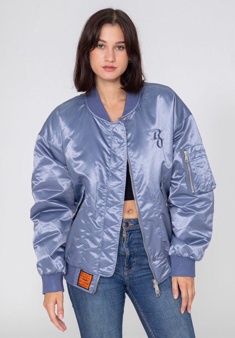 Jasnoniebieska satynowa kurtka bomberka z prążkowanymi mankietami, zamkiem błyskawicznym na rękawie oraz pomarańczowym detalem metki. Model nosi ją z dżinsami.