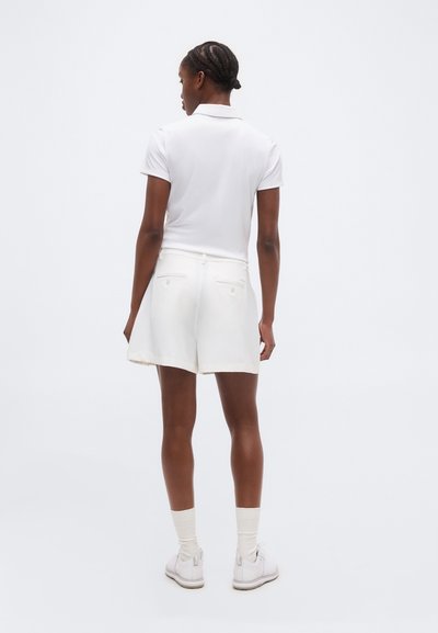 Γυναίκα που φοράει λευκό πόλο μπλουζάκι με κοντά μανίκια, λευκό σορτς με πίσω τσέπες, λευκές κάλτσες και λευκά αθλητικά παπούτσια, όρθια και με την πλάτη στραμμένη προς τα εμπρός.