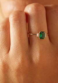 Goldring mit einem großen smaragdschliff grünen Edelstein, flankiert von zwei kleinen rechteckigen klaren Steinen an einem Finger einer Person.
