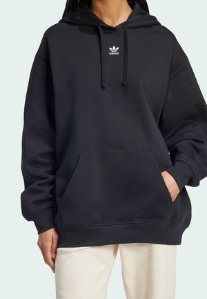 Osoba ubrana w oversize czarną bluzę Adidas z kieszenią z przodu i białe spodnie, stojąca na jasnym tle.