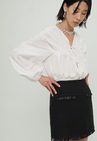 Blouse blanche à manches longues avec décolleté en V et détail à lacets, associée à une mini-jupe noire texturée avec des accents de franges et des poches.