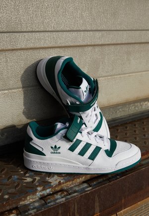 Białe i zielone wysokie trampki Adidas ułożone na zardzewiałej metalowej powierzchni na tle beżowej, teksturowanej ściany.