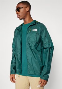 Groene lichte ritsjack met een capuchon en logo op de linkerkant van de borst. Draagt over een blauwe T-shirt, gecombineerd met beige broeken.