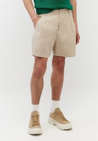 Bermuda beige in lino con design sartoriale, dotati di tasche laterali e frontale piatto. Indossati con sneakers beige e calzini bianchi.