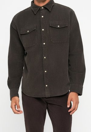 Homme portant une chemise à manches longues en velours côtelé marron foncé avec des poches poitrine boutonnées et un pantalon en velours côtelé marron foncé assorti.