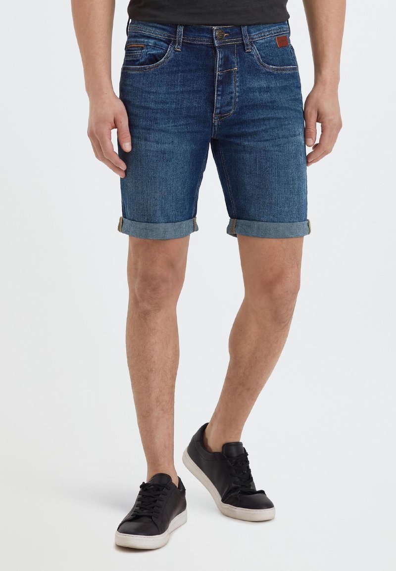 Mann trägt hochgekrempelte dunkelblaue Jeansshorts und schwarze Sneakers und steht mit leicht gespreizten Beinen vor einem schlichten weißen Hintergrund.