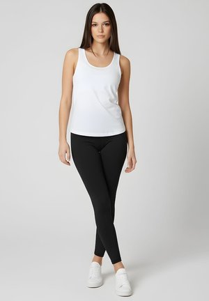 Joven con largo cabello castaño, vistiendo una camiseta blanca sin mangas, leggings negros y zapatillas blancas, de pie frente a un fondo gris claro.