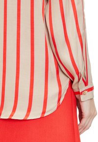 Long-sleeve blouse in beige fabric with bold red vertical stripes and buttoned cuff detail, paired with a solid red skirt.