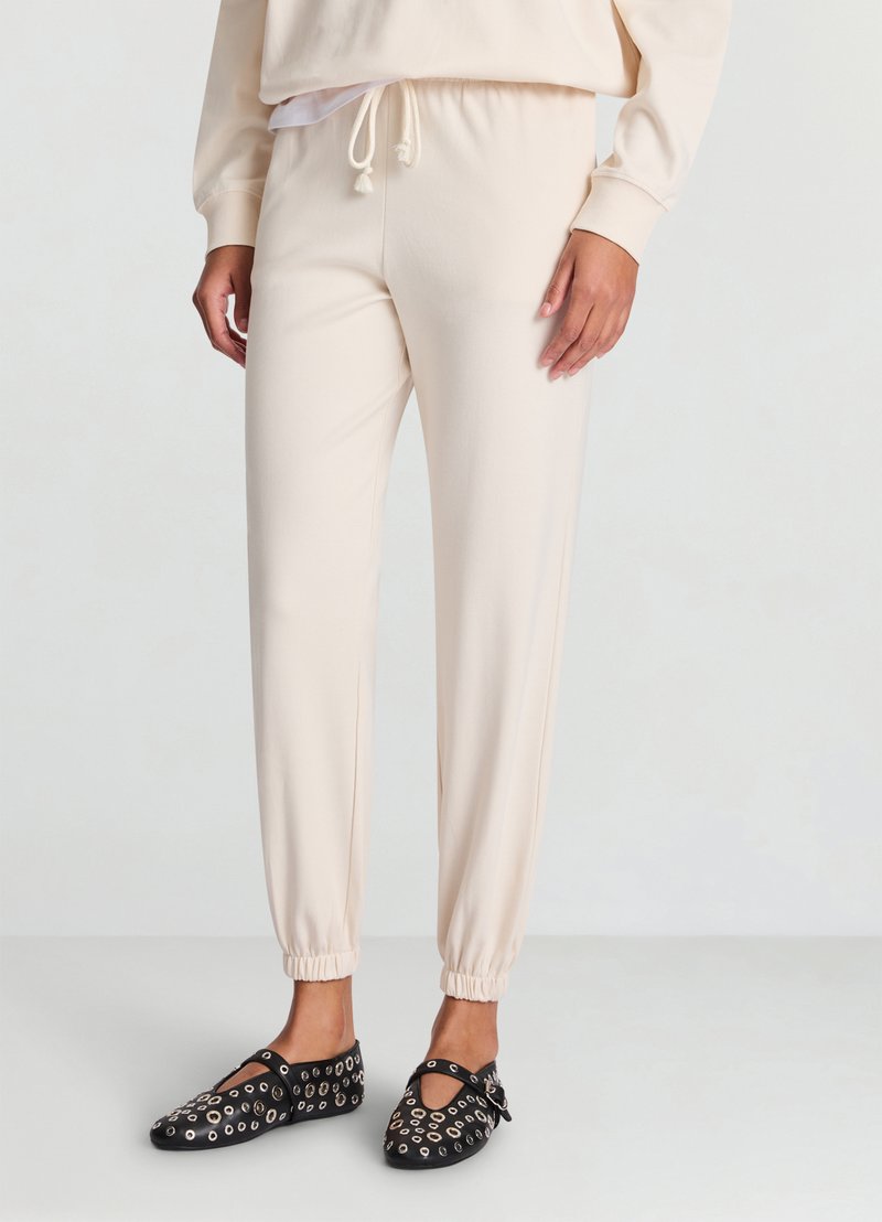 Pantalon de jogging crème clair avec taille élastique, chevilles resserrées et texture lisse, associé à des chaussures noires avec œillets métalliques.