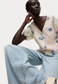 Blusa floreale trasparente con motivi blu e viola, abbinata a jeans a gamba larga azzurri. Texture morbide e design rilassato.