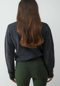 Dunkelgrauer, strukturierter Strickpullover mit gerippten Bündchen, kombiniert mit eng anliegenden, dunkelgrünen Hosen mit seitlichen Taschen, von hinten betrachtet.