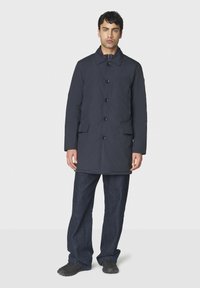 Marineblauer Hemdengartenmantel mit zwei Vordertaschen und Kragen, aus glattem Stoff. Kombination mit dunklen Jeans und schwarzen Schuhen.