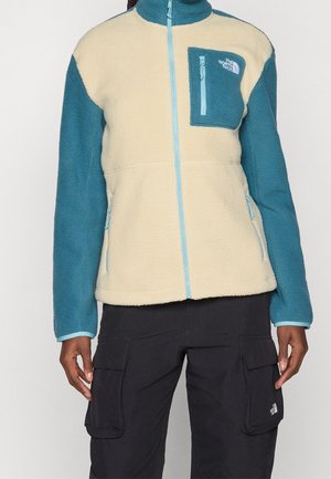 Osoba ubrana w dwukolorową polarową kurtkę z niebieskimi rękawami i kremowym tułowiem, z kieszenią na piersi na zamek, w połączeniu z czarnymi spodniami cargo.