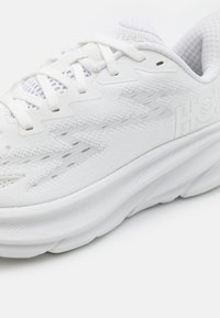 Vit sportskor med ett strukturerat meshupp, utrustad med en tjock sula, platta snören och minimalistisk branding. Slät yta och rund tådesign.