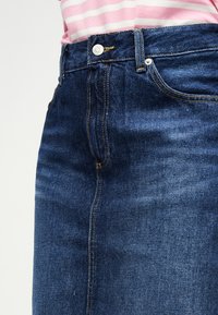 Κοντινό πλάνο μπλε τζιν φούστας με κίτρινα ραφές, που φοριέται με ροζ και λευκή ριγέ μπλούζα.