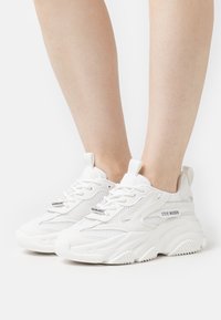 Chaussures de sport blanches avec tige en mesh et synthétique, semelle épaisse, col rembourré et lacets plats. Nom de la marque affiché sur le côté.