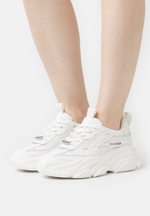Chaussures de sport blanches avec tige en mesh et synthétique, semelle épaisse, col rembourré et lacets plats. Nom de la marque affiché sur le côté.