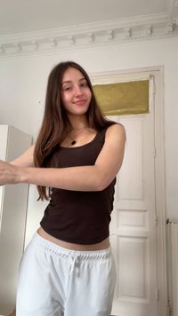 Jeune femme aux longs cheveux bruns, portant un débardeur foncé et un pantalon blanc, se tient à l'intérieur près d'une porte blanche avec un rideau doré au-dessus.