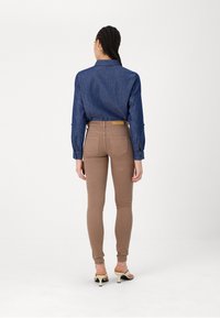 Chemise en denim à manches longues de couleur bleu foncé, associée à un pantalon slim marron, présentant des poches arrière et un design minimaliste. Sandales à talons hauts visibles.