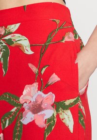 Tecido vermelho com grandes padrões florais em rosa e folhas verdes. O design inclui bolsos visíveis de lado.