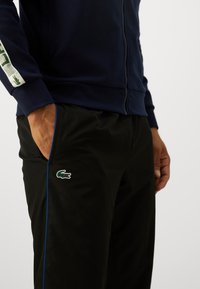 Jachetă bleumarin cu fermoar și finisaj texturat, purtată cu pantaloni negri care au o dungi albastre și un logo verde de crocodil pe piciorul drept.