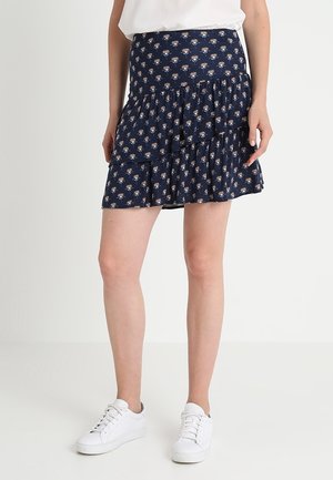 Kobieta ubrana w granatową mini spódnicę w kwiaty z falbanami, w białe trampki i biały top, stojąca na tle jednolitego jasnego tła.