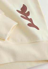 Sweat-shirt de couleur crème avec texture côtelée, orné d'un détail appliqué en forme de feuille marron sur le devant. Finition lisse et design décontracté.