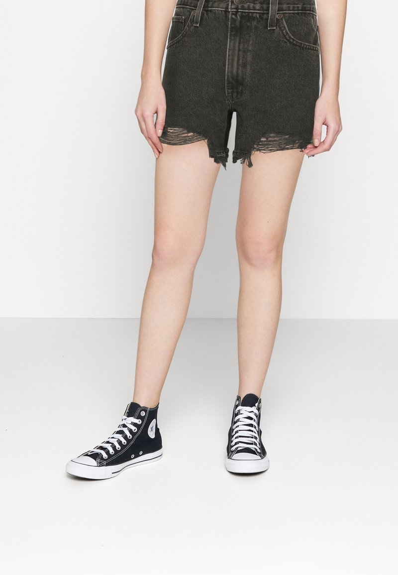 Personne portant des baskets montantes noires et un short en jean noir déchiré, debout sur un sol blanc uni, devant un fond blanc.