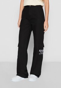 Black cargo pants made of cotton with side pockets. Features a logo patch on the left leg and a relaxed, wide-leg fit. Paired with white sneakers.