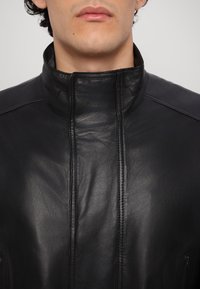 Musta nahkatakki, jossa on korkea kaulus, sileä pinta ja näkyvät ompeluyksityiskohdat. Varustettu vetoketjullisilla sivutaskuilla ja minimalistisella muotoilulla.