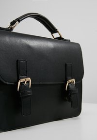 Borsa porta documenti nera in pelle con chiusura a patta, con hardware color oro e due cinghie con fibbia. Texture liscia e forma strutturata.