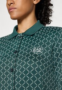 Primo piano di una persona che indossa una camicia a bottoni verde con motivo e il logo EA7 Emporio Armani sul petto.