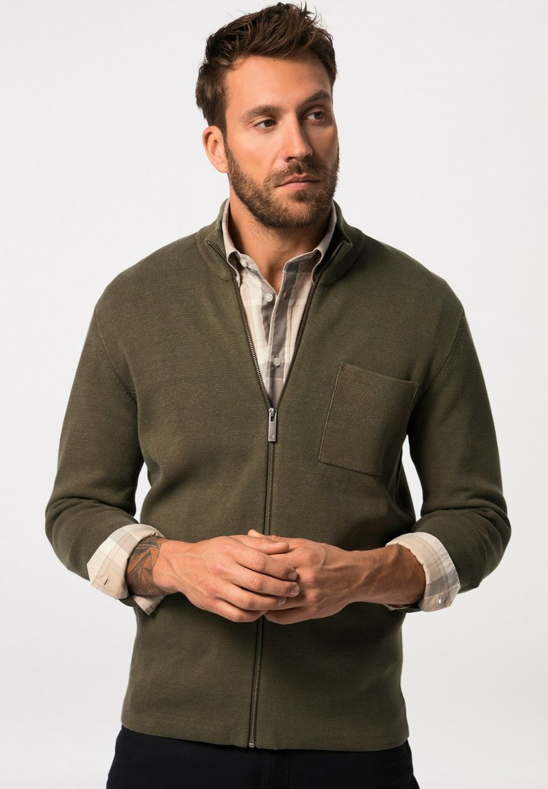 Chaqueta de forro polar verde olivo con cremallera y un bolsillo en el pecho, que presenta una textura de punto y puños de cuadros enrollados, combinada con una camisa a cuadros.