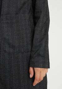 Tissu à motif chevrons noirs avec détails blancs. Gros plan sur une manche montrant le design texturé et une main reposant sur le vêtement.